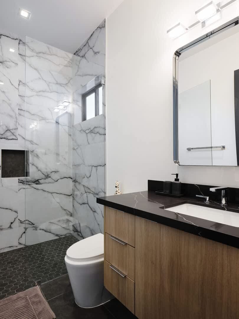 A modern bathroom with a marble wall, a sink, a toilet, and a mirror.