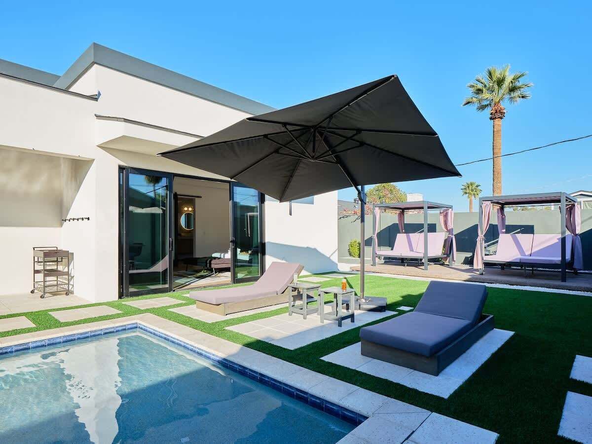 Un área de piscina moderna al aire libre con sillones de descanso, una sombrilla grande y una entrada con puerta de vidrio.