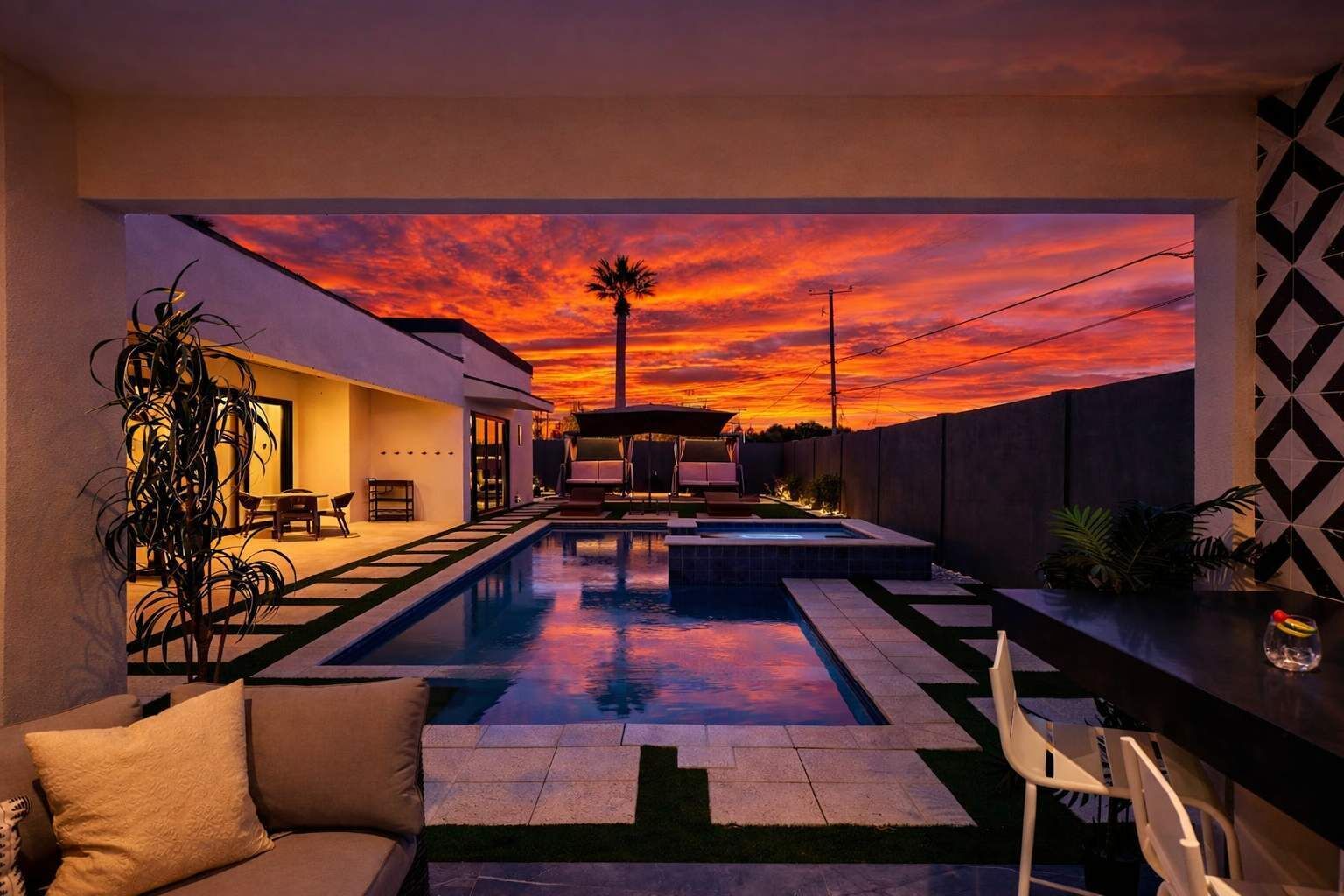 A modern outdoor pool area with a sunset view, a patio with seating, and a geometric wall design.
