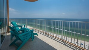 A balcony with a view of the ocean, equipped with two turquoise Adirondack chairs and a glass door leading to the interior. - Phoenix Gulf Towers 1908 4 Bedroom Condo (Orange Beach)
