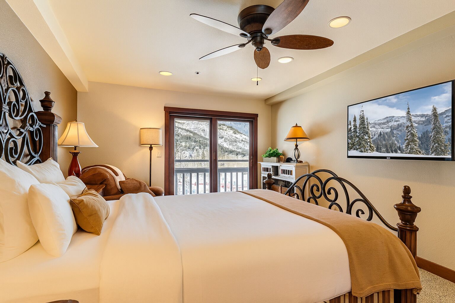 A hotel room with a large bed, a ceiling fan, a balcony with a view of snow-covered mountains, and a television on the wall.