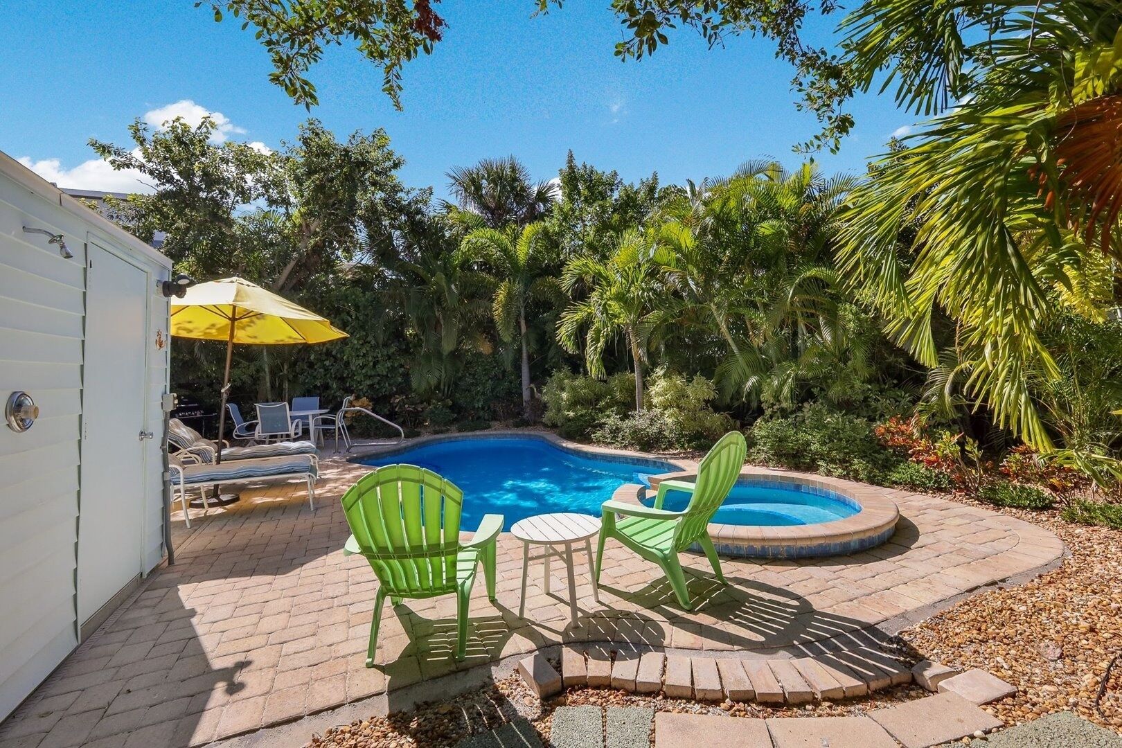A backyard with a swimming pool, green chairs, a yellow umbrella, and a white building.