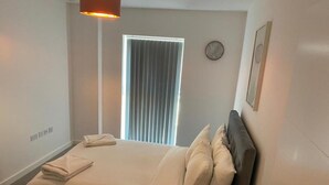 Una cama bien hecha con almohadas y una toalla doblada, una lámpara de noche, una ventana con persianas y un reloj en la pared.