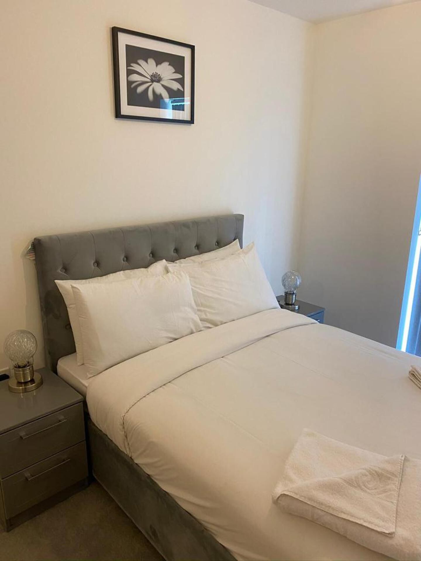 A neatly made bed with white linens, a grey upholstered headboard, and a bedside table with a lamp.