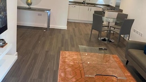 Una sala de estar moderna con una mesa de centro de vidrio, un sofá y un comedor con una mesa y sillas de vidrio. Se ve una cocina con gabinetes blancos y electrodomésticos de acero inoxidable al fondo.