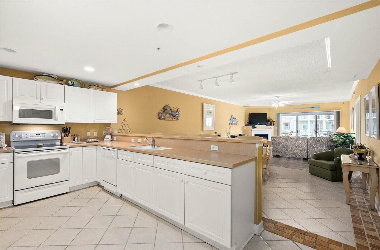 A modern kitchen with white cabinets, a built-in oven, and a microwave. There is a dining area with a sofa and a small table.