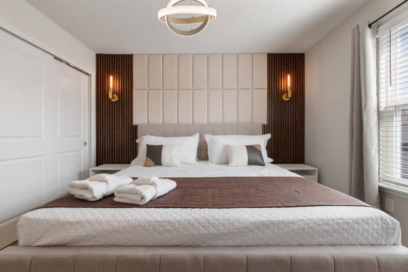A modern bedroom with a large bed, two bedside tables, a headboard with built-in lighting, and a window with blinds.