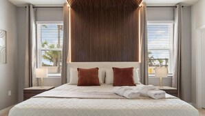 A large bed with a quilted white cover, two brown pillows, and a wooden headboard. There are two bedside tables with lamps and a view of palm trees through the windows. - New Vacation Home Windsor Island by Disney w/Pool (Davenport)