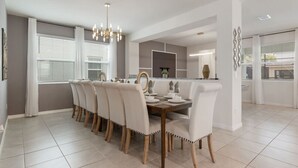 Un espace repas avec une longue table et des chaises, un lustre et une cuisine en arrière-plan.