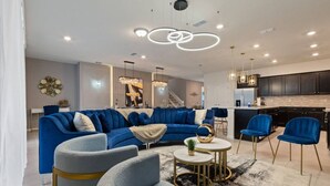 Un salon moderne avec un canapé bleu, des fauteuils gris, une table basse ronde et un coin cuisine doté de meubles sombres et de chaises bleues.