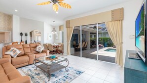 A spacious living room with a sofa, coffee table, and a ceiling fan. There is a dining area with chairs and a table, and a TV on a stand. - Stunning Briarwood Lakefront Pool and Spa Home (Naples)