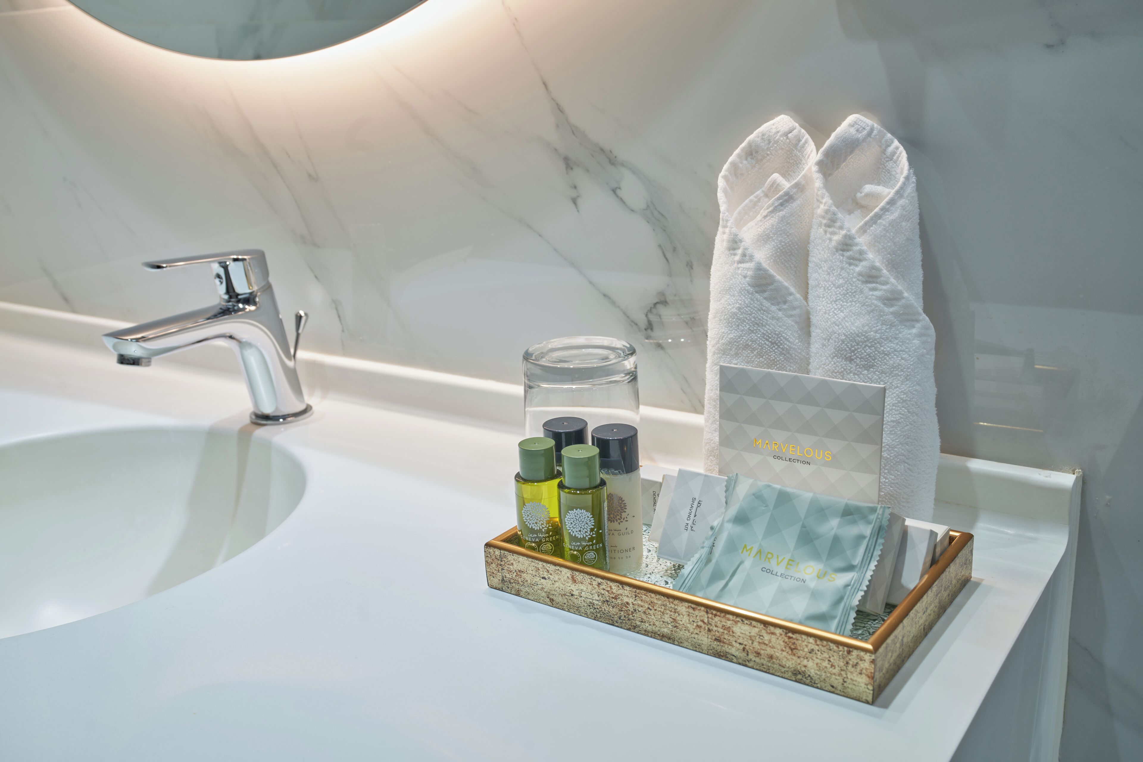 A bathroom sink with a marble wall, a modern faucet, and a tray holding toiletries and a towel.