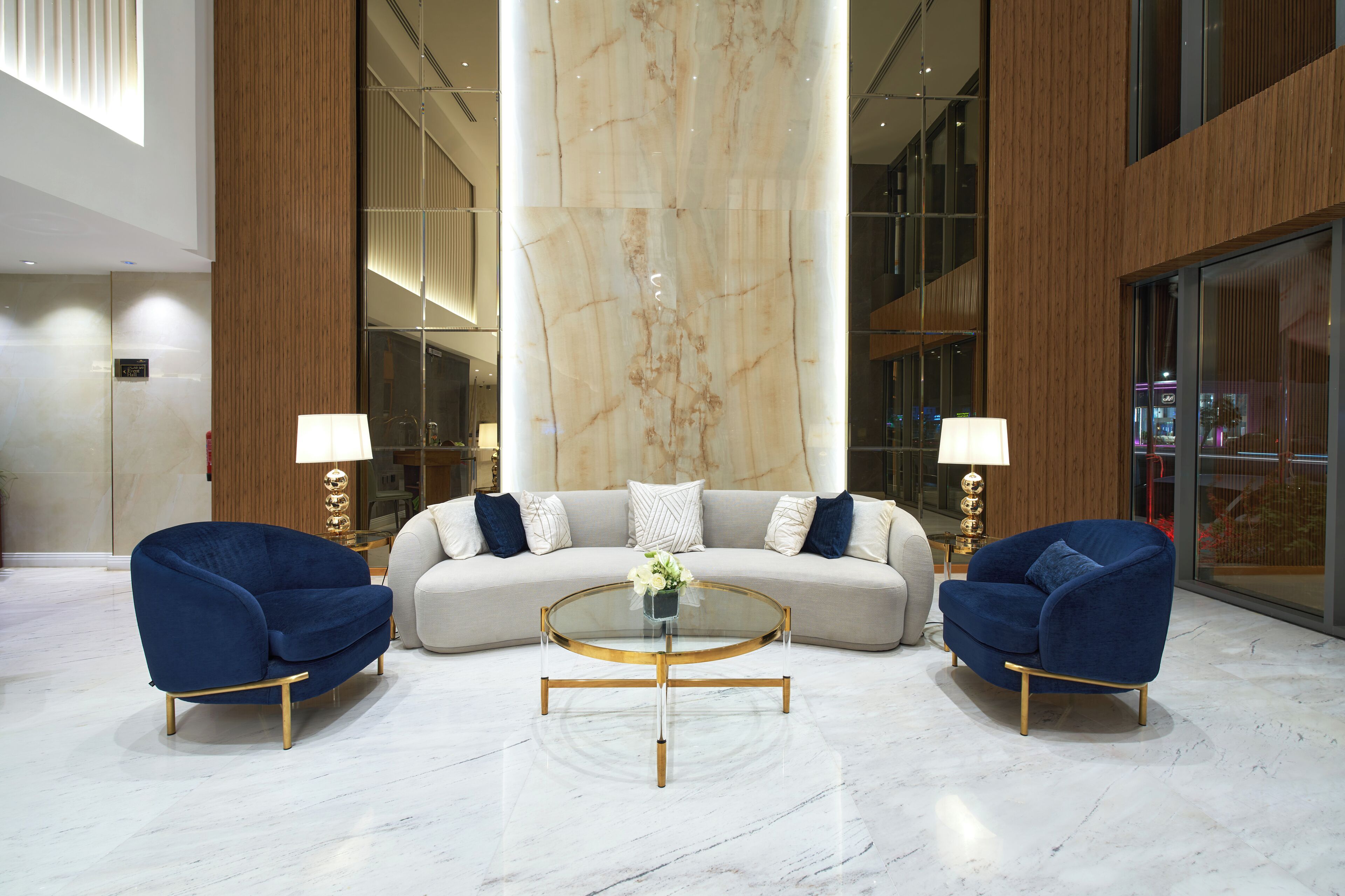 A modern lobby with a large marble column, a round glass coffee table, two blue armchairs, and a beige sofa with white and blue pillows.