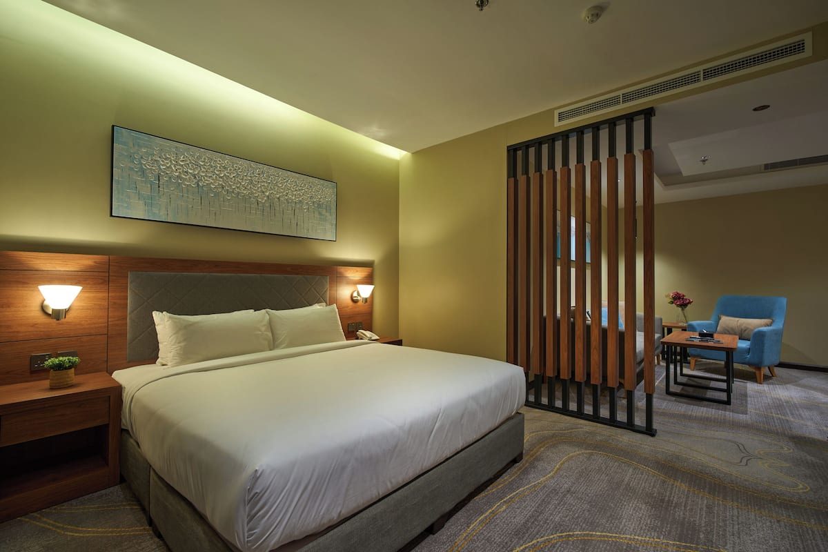 A hotel room with a large bed, bedside lamps, a wall-mounted artwork, a wooden partition, a blue armchair, and a small table.