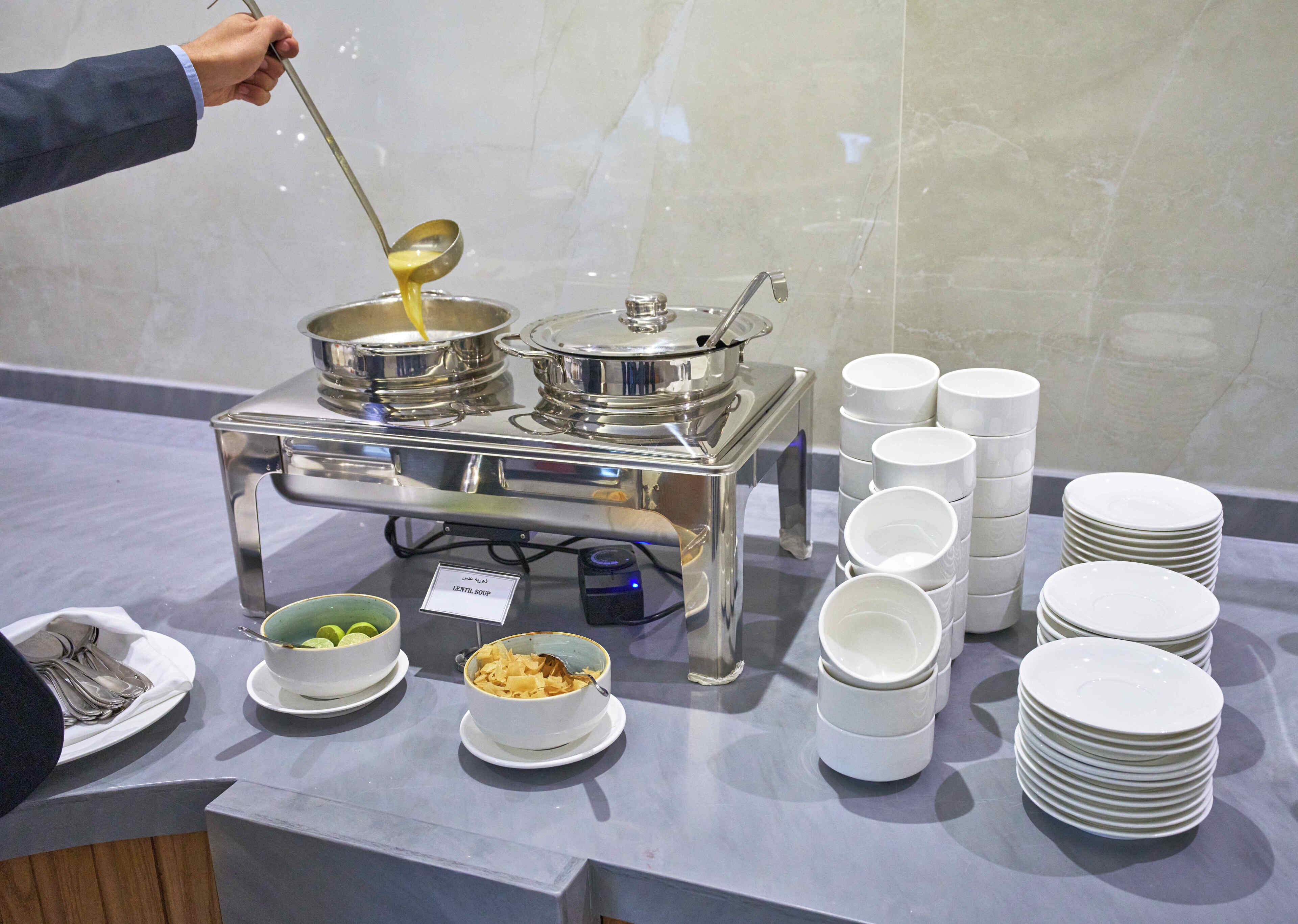 Montaje de bufé con varios recipientes de comida, platos apilados y un cartel que indica 'LENTIL SOUP'.