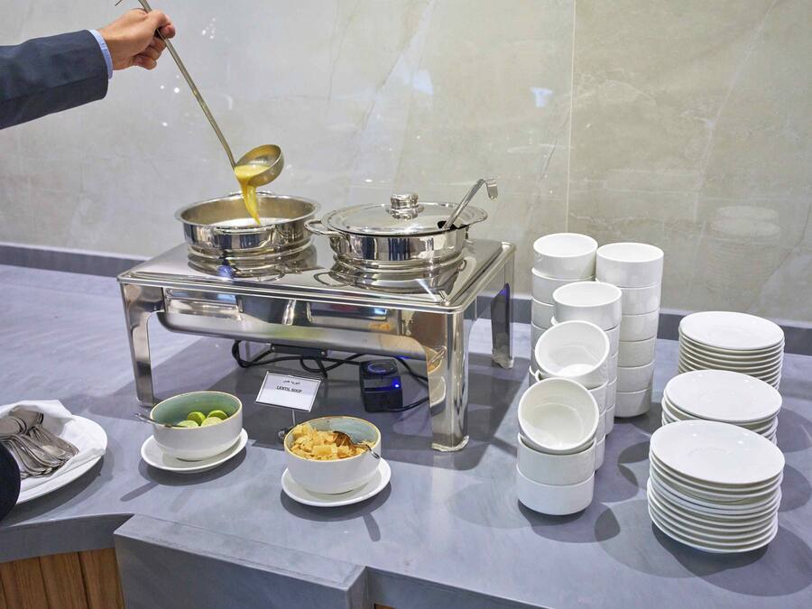 Montaje de bufé con varios recipientes de comida, platos apilados y un cartel que indica 'LENTIL SOUP'.