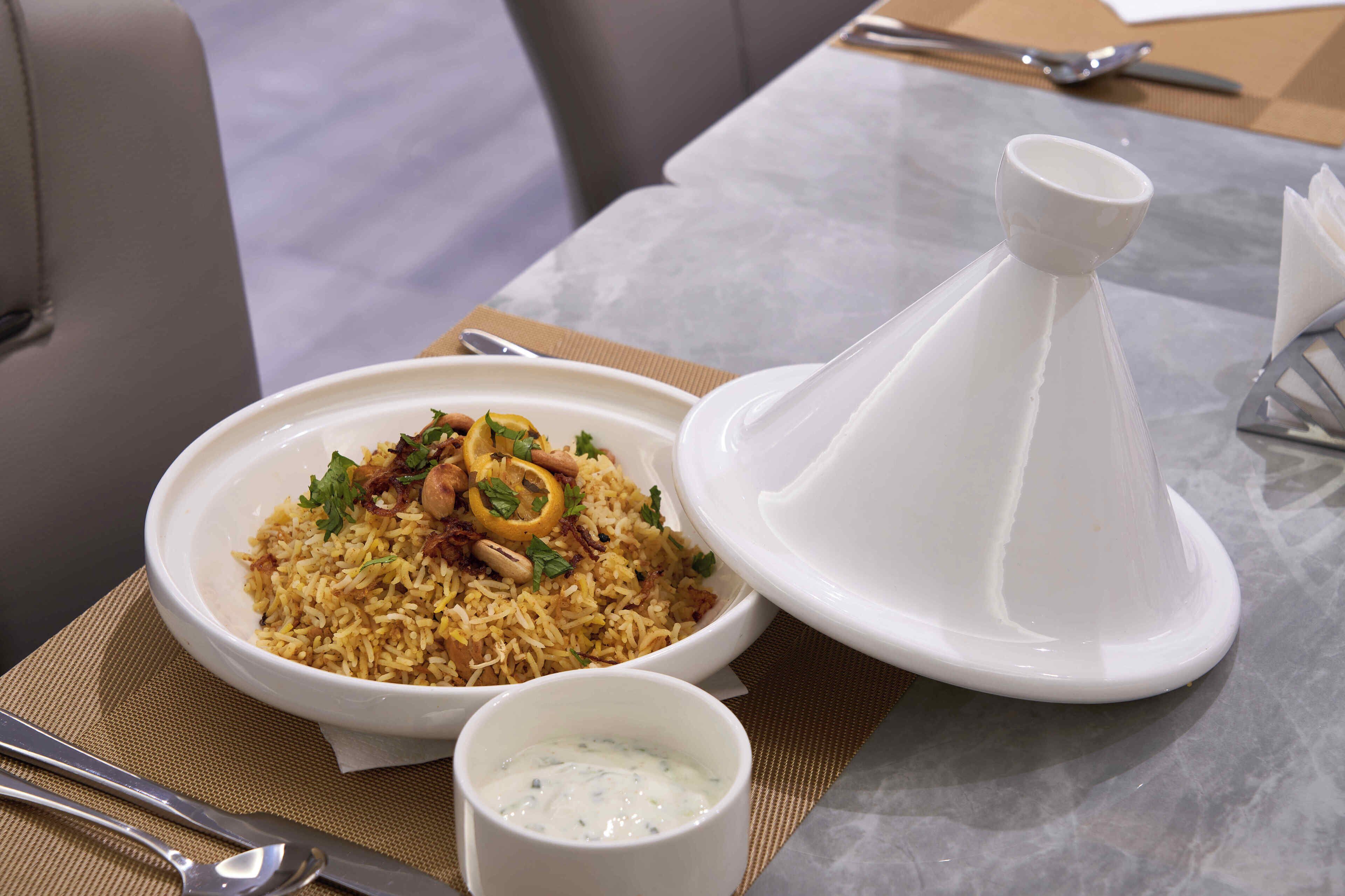 A table with a bowl of biryani, a white conical serving dish, and a small bowl of raita.