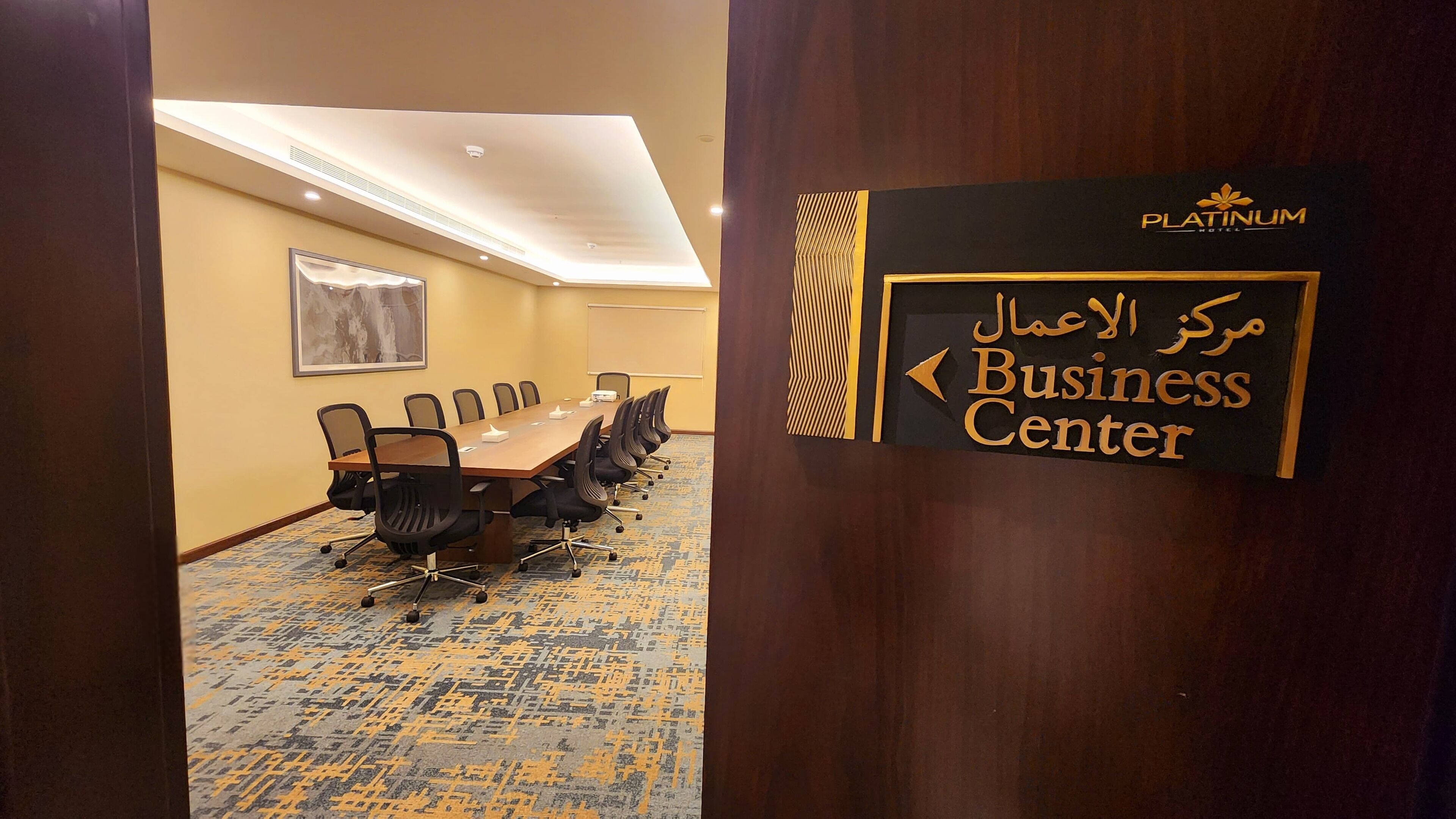 Une salle de conférence avec une longue table et des chaises, un panneau indiquant le centre d’affaires, et un tapis à motifs.