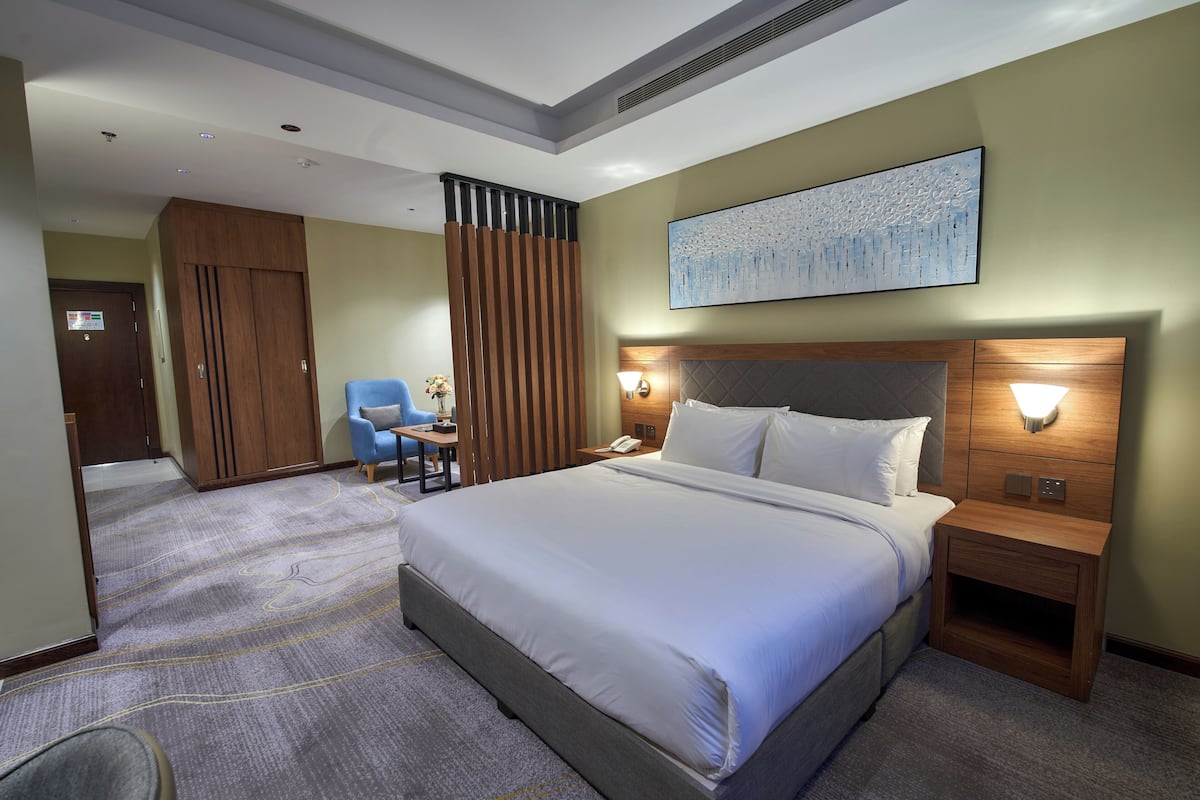 A hotel room with a large bed, a desk with a chair, a wooden wardrobe, a wall-mounted artwork, and a nightstand with a lamp.