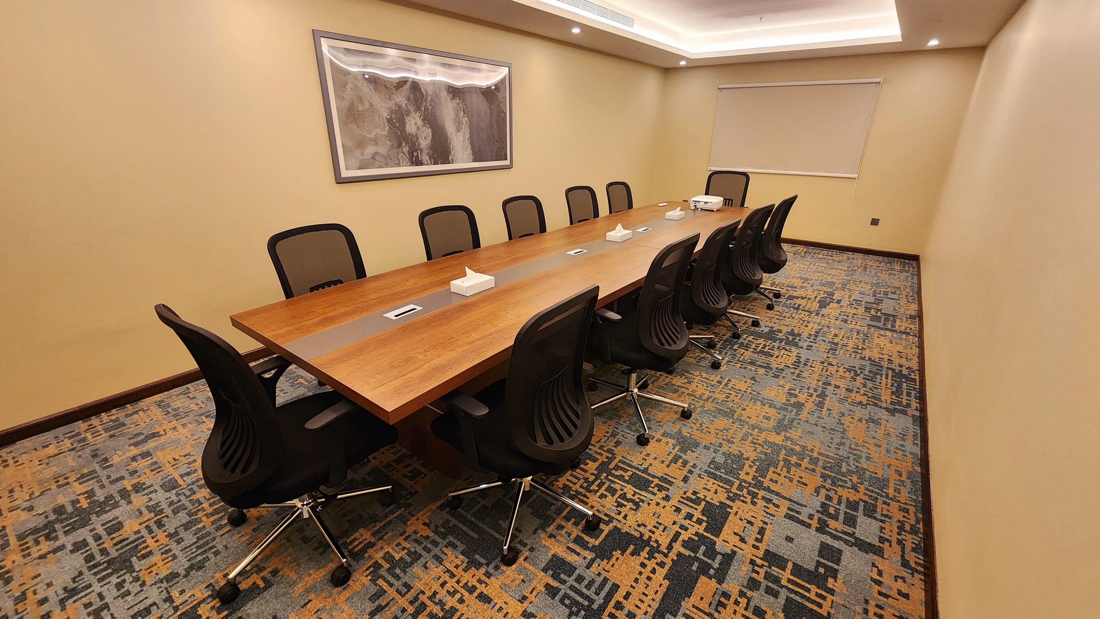 Uma sala de conferências com uma mesa comprida, cadeiras pretas, um projetor e piso coberto de carpete.
