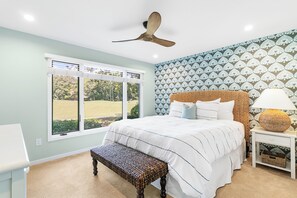 A bedroom with a large bed, a ceiling fan, a window with a view of a lawn, and a patterned wall. - Updated villa on golf course, 3 min to beach! (Kiawah Island)