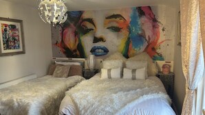A bedroom with two beds, fluffy white bedding, a colorful abstract mural on the wall, a chandelier, and a small side table with a vase. - Immaculate 3-bed House in Bridgnorth (Bridgnorth)