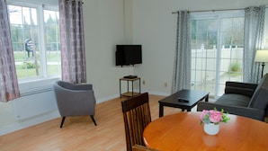 A living room with a wooden dining table, two chairs, a sofa, a television, and a view of a backyard through a sliding glass door. - Prospectors Inn (Kirkland Lake)