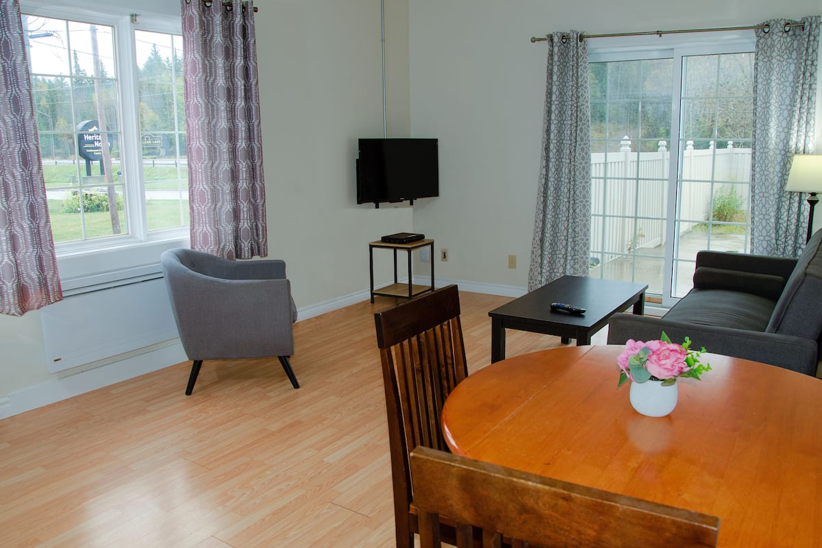 Un salon comprenant une table à manger en bois, deux chaises, un canapé, un téléviseur et une vue sur un jardin à travers une porte-fenêtre coulissante.