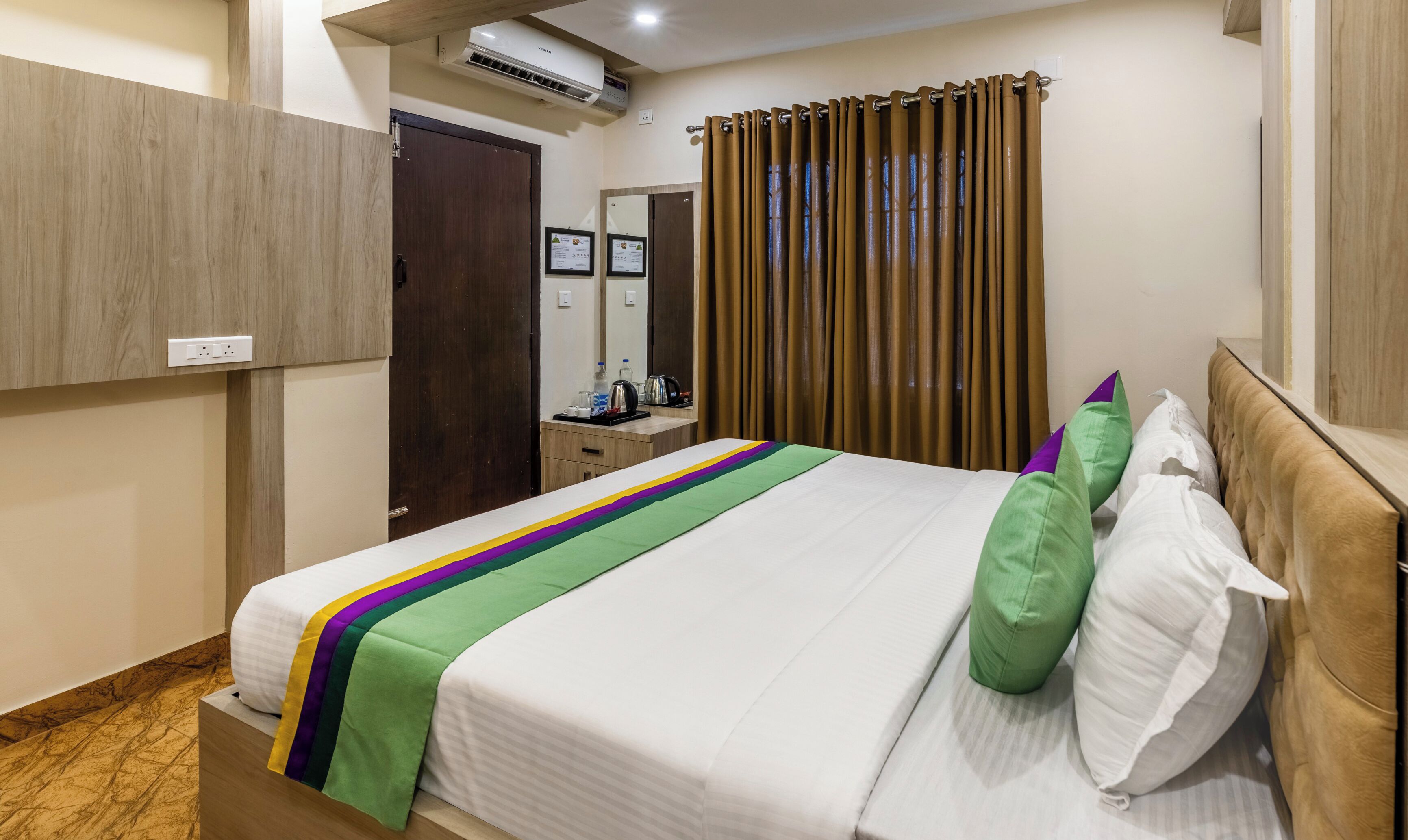 Habitación de hotel con cama, cabecera de madera, mesita de noche, espejo y ventana con cortinas.