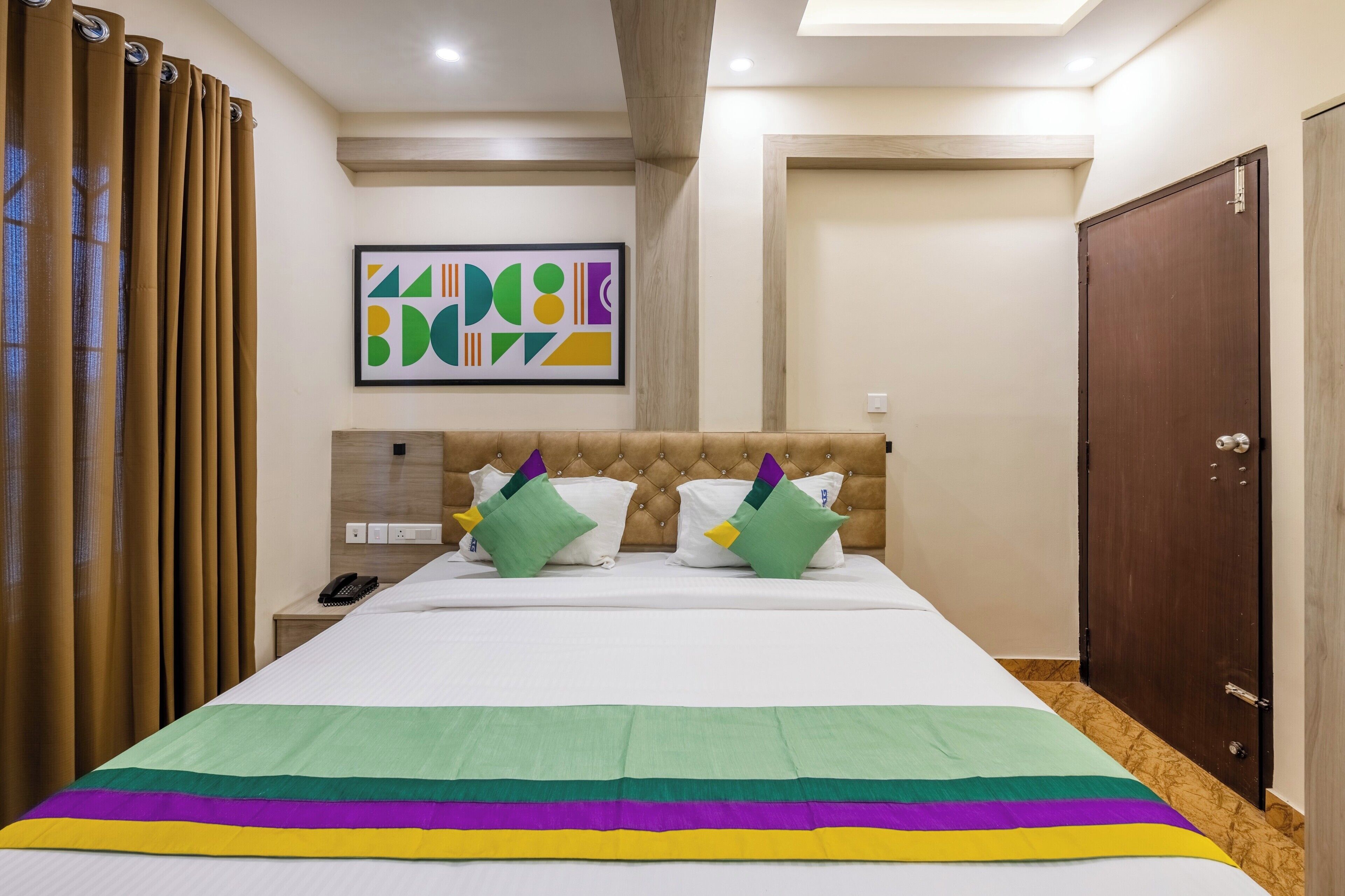 Habitación de hotel con una cama grande, cabecera, mesita de noche con teléfono, puerta y un muro con un cuadro abstracto de colores.