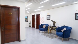 A spacious lobby with a wooden door labeled "102", a blue armchair, a small table with decorative items, and a framed picture on the wall. - Hotel Elegant Executive Suites (Multan)