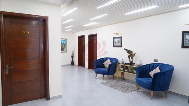 Eine geräumige Lobby mit einer Holztür mit der Aufschrift „102“, einem blauen Sessel, einem kleinen Tisch mit Dekoration und einem gerahmten Bild an der Wand.