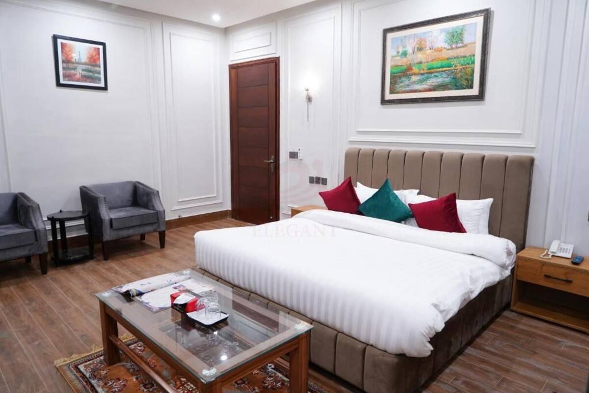 A hotel room with a large bed, two armchairs, a small table, a wooden nightstand, and a painting on the wall.