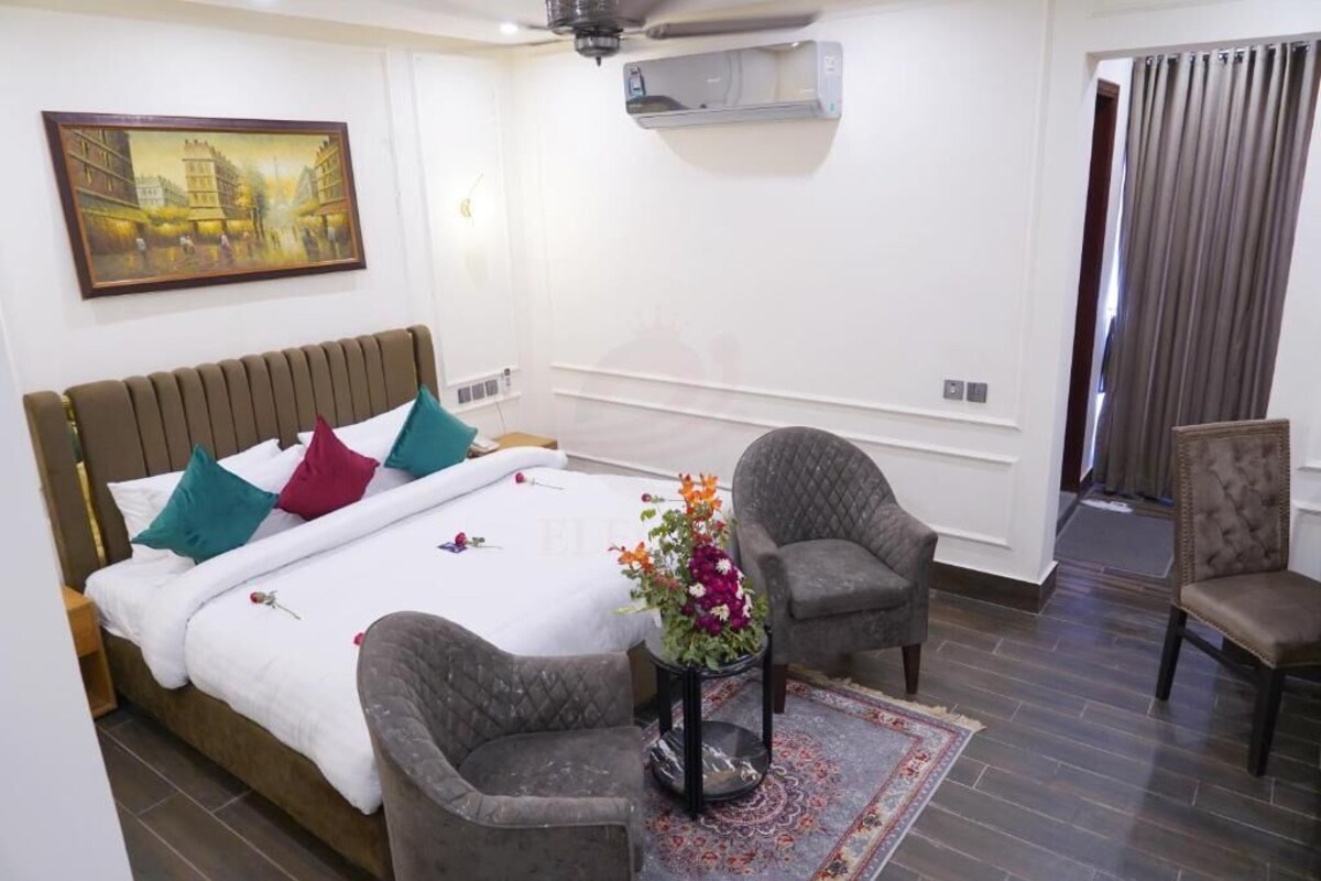 A hotel room with a bed, two armchairs, a small table with a vase of flowers, a painting on the wall, and an air conditioning unit.
