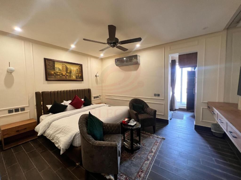Un dormitorio con una cama, dos sillones, una mesita, un ventilador de techo, un aire acondicionado y un cuadro en la pared.