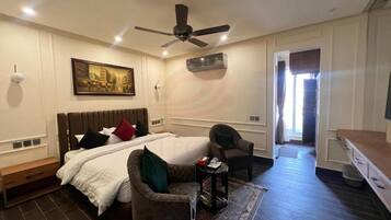 Un dormitorio con una cama, dos sillones, una mesita, un ventilador de techo, un aire acondicionado y un cuadro en la pared.