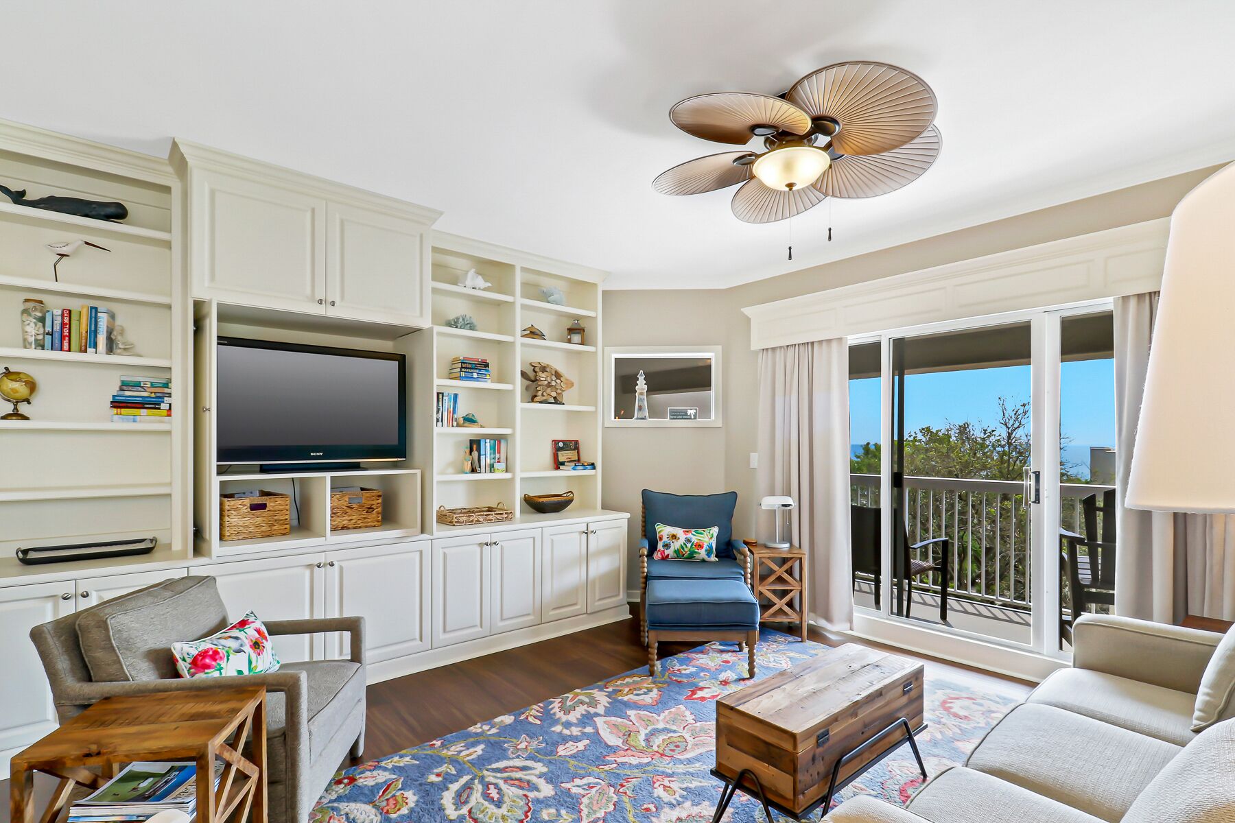 A spacious living room with a flat-screen TV, built-in shelves, a blue armchair, a wooden coffee table, and a balcony with a view.