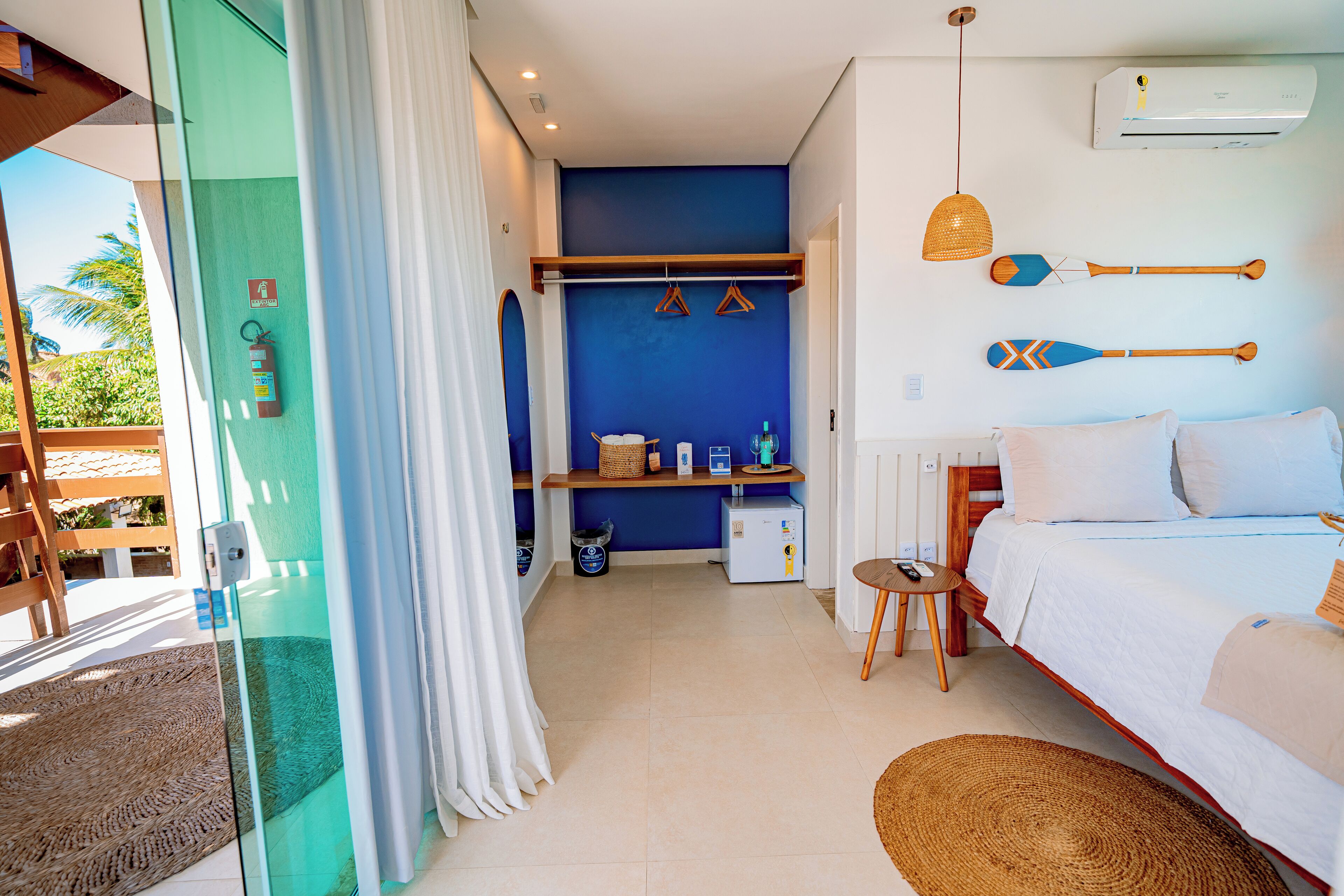 A hotel room with a bed, a small table, a chair, a shelf with items, and a view of the outdoors.