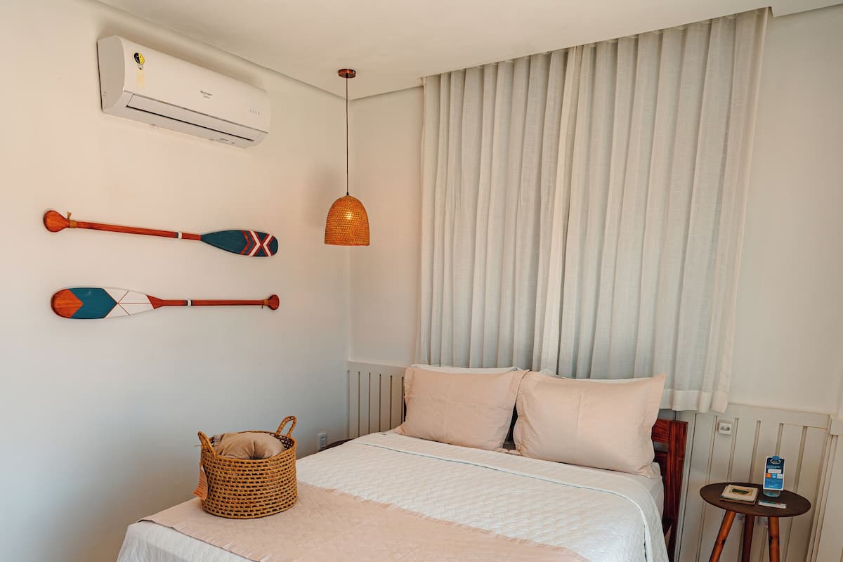 A neatly arranged bedroom with a large bed, a bedside table, a wall-mounted air conditioner, and a small table with a lamp.