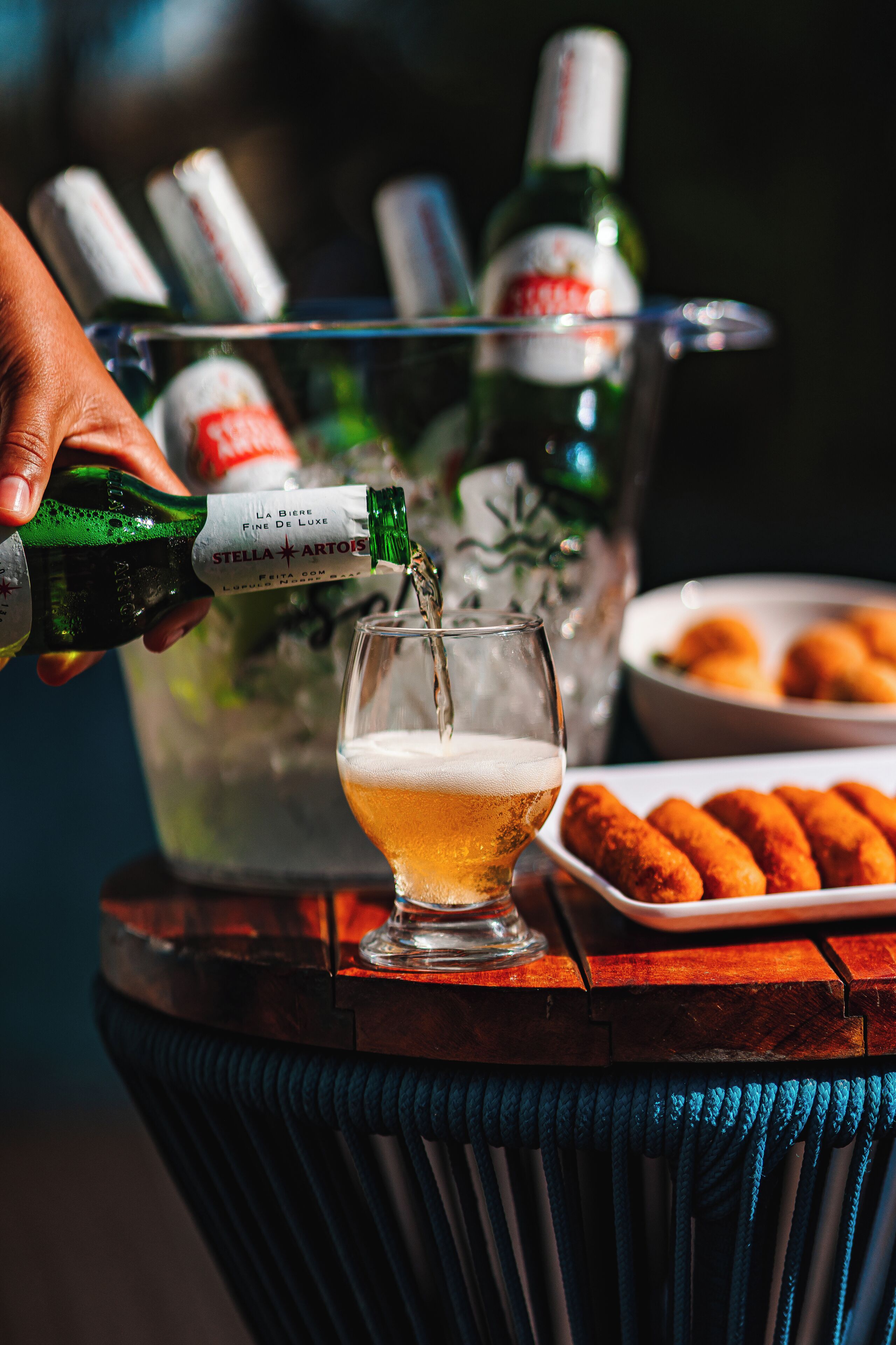 Una mano sirve de una botella a un vaso sobre una mesa de madera, con un bol de comida y una botella de Stella Artois al fondo.