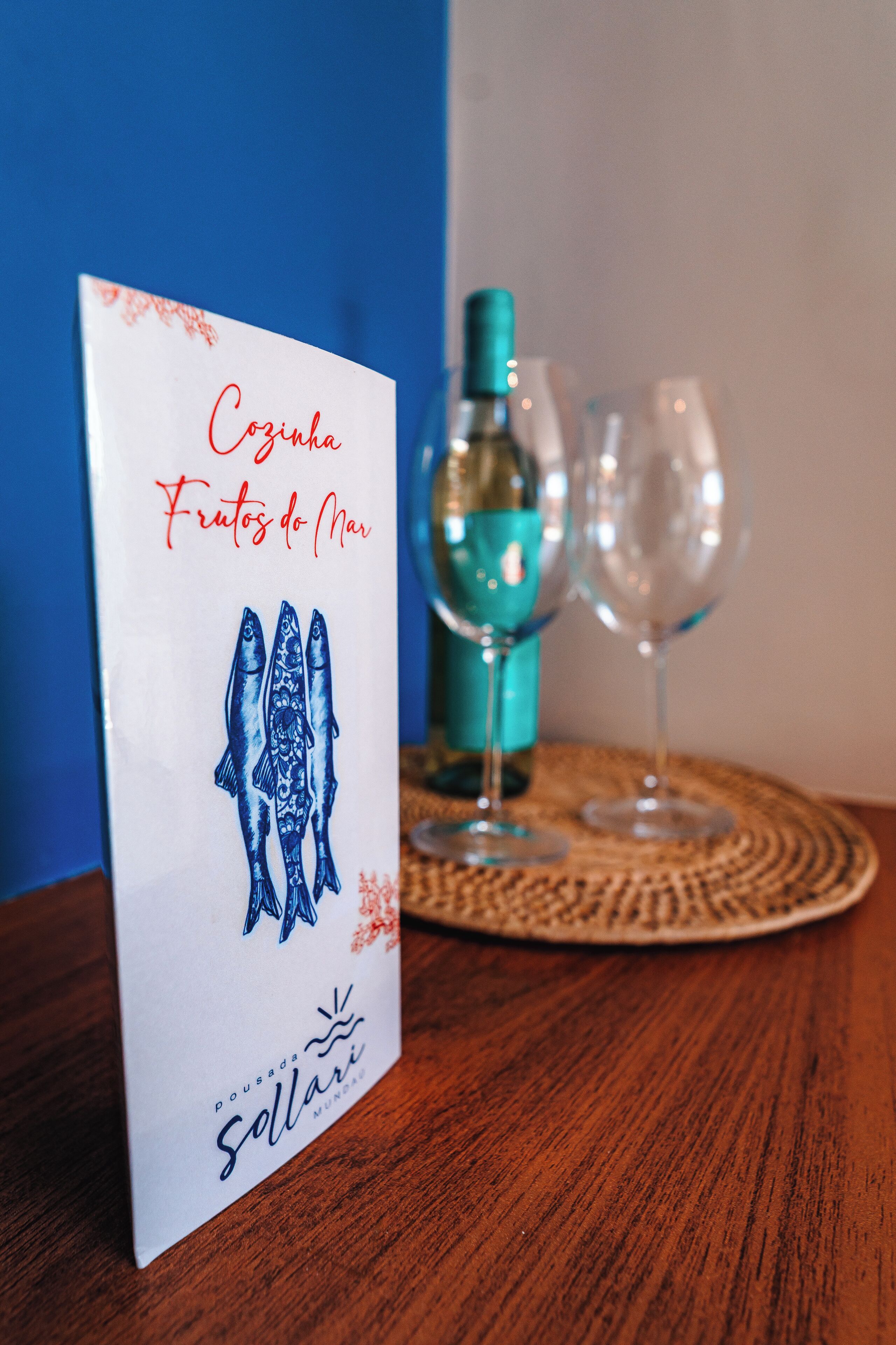 Eine Speisekarte mit der Aufschrift "Cozinha Frutos do Mar" und einer Illustration von Meeresfrüchten, platziert auf einem Holztisch zusammen mit zwei Weingläsern und einer Flasche.