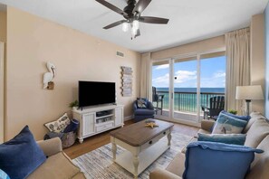 A living room with a flat-screen TV, a ceiling fan, and a view of the ocean through large sliding glass doors.