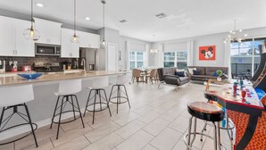 A modern kitchen with white cabinets, a central island with stools, a dining area with a sofa, and a colorful arcade table.