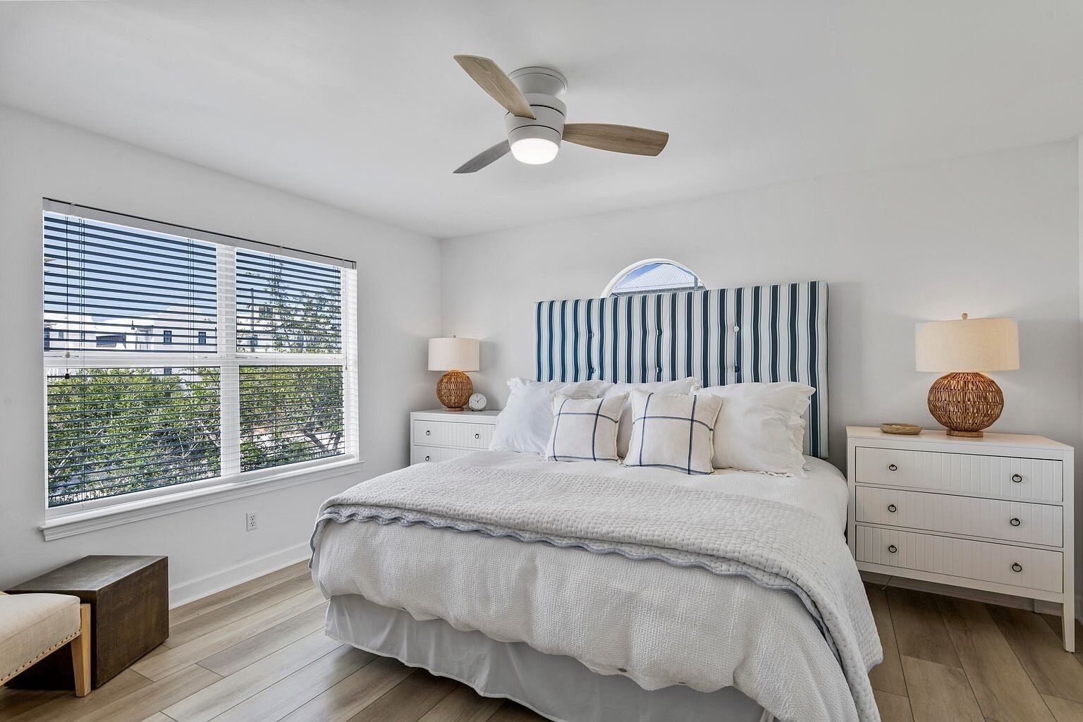 A bedroom with a bed, two bedside tables, a ceiling fan, a window with blinds, and a view of greenery outside.