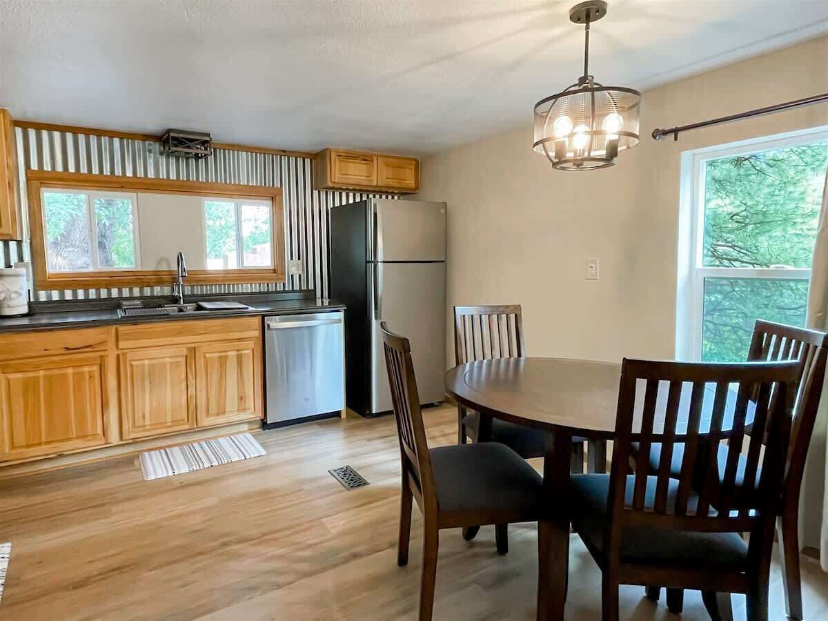 Une cuisine avec des meubles en bois, un réfrigérateur en acier inoxydable, une table à manger avec des chaises et une fenêtre donnant sur des arbres.