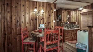 A rustic wooden interior with a round dining table, two red chairs, a small kitchen area with a refrigerator, and a wall-mounted clock. - Lofty Pines Cabin (Ruidoso)