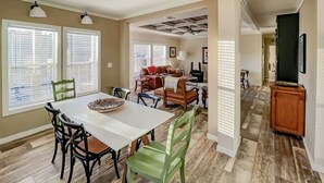 A dining area with a white table, chairs, and a pizza on the table. There are two windows with blinds, a ceiling fan, and a living area with a couch and chairs.