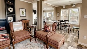 A spacious living area with a dining table, two wooden chairs, a patterned rug, and a kitchen in the background.
