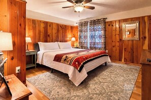 Ein Schlafzimmer mit Holzwandverkleidung, einem Bett, zwei Nachttischlampen, einem Deckenventilator und einem gemusterten Teppich.