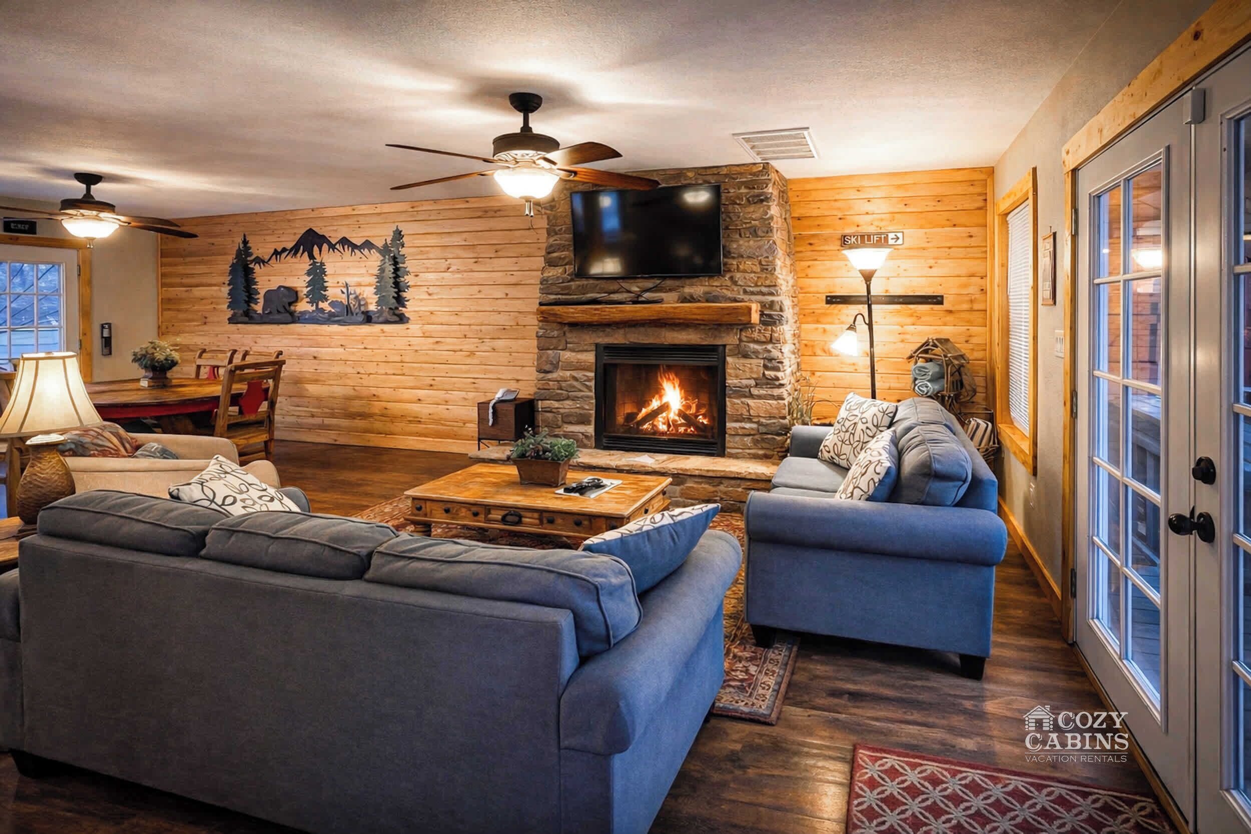A cozy cabin interior with a stone fireplace, two sofas, a television, and a ceiling fan.